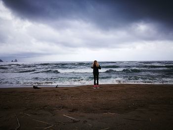Rear view of woman standing at shore against cloudy sky