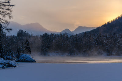 Frozen lake by trees against sky during sunrise