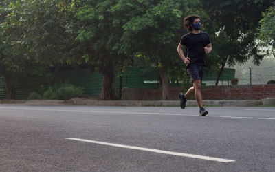 Side view of a man running while wearing a mask on the road with trees in the background.