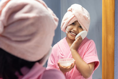 A cute indian girl child in pink bathrobe applying powder in front of mirror