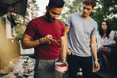 Young man photographing cherries with mobile phone during social gathering