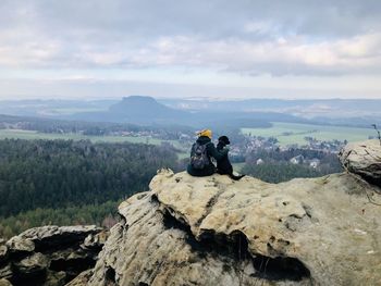 Rear view of woman sitting with dog on rock against sky