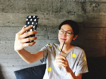 Smiling woman taking selfie while having coffee against wall