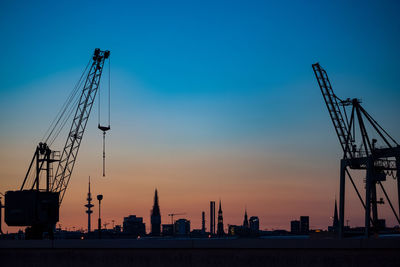 Silhouette cranes at commercial dock in front of city silhouette against sky during sunset