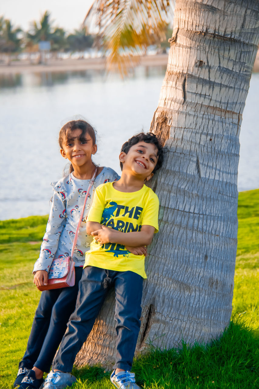 child, childhood, two people, plant, togetherness, smiling, men, full length, happiness, emotion, women, nature, female, grass, tree, family, person, yellow, adult, portrait, positive emotion, spring, love, looking at camera, cheerful, day, casual clothing, bonding, leisure activity, water, jeans, footwear, clothing, outdoors, toddler, standing, lifestyles, fun, tree trunk, enjoyment, one parent, cute