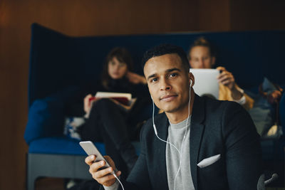 Portrait of confident mid adult businessman listening to in-ear headphones from mobile phone sitting against colleagues