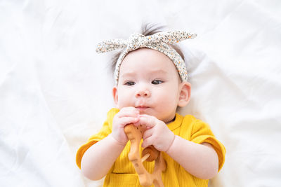 Healthy asian baby girl in yellow bodysuit plays with wooden toy teether for teeth on white bedding