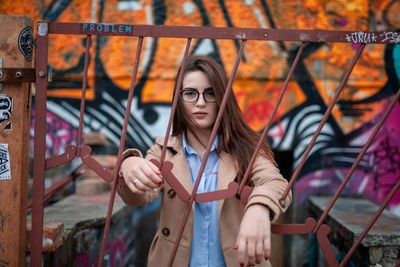 Portrait of young woman standing behind metal gate against graffiti wall