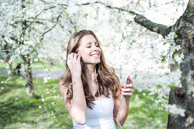 Young woman, closed eyes, holding phone, listening to music via earphones under the blooming trees