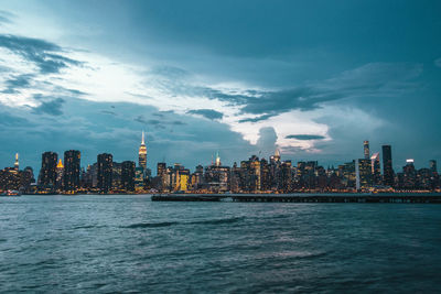 Sea by modern buildings in city against sky at dusk