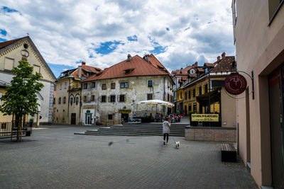 Rear view of woman walking with dog on walkway in town against cloudy sky