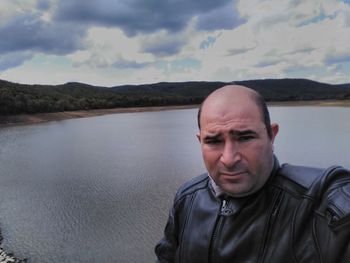 Portrait of mature man standing by lake against cloudy sky