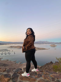 Portrait of young woman standing at beach against sky during sunset