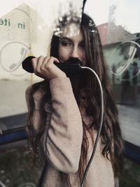 Woman talking on pay phone while standing at booth seen through glass