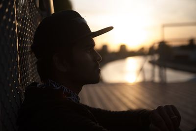 Profile view of young man sitting by fence during sunset