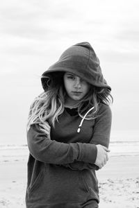 Portrait of woman with arms crossed wearing hooded shirt at beach against sky