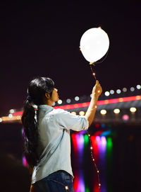 Side view of woman holding helium balloon against sky at night