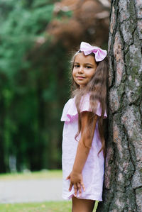 Portrait of a cute little girl leaning against a big tree with her back in the forest in summer