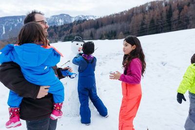 Father with daughters stand next to snowman they made in the mountains