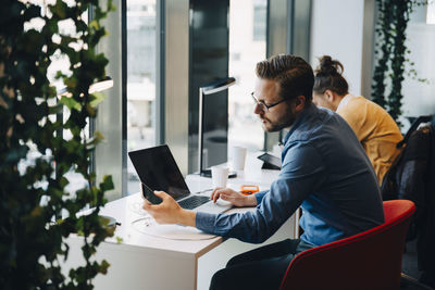 Side view of businessman using mobile phone and laptop while sitting by colleague at desk in office