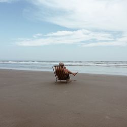 Man sitting on deck chair at beach against sky