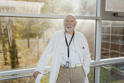 Portrait of senior male doctor leaning on railing against window at hospital corridor