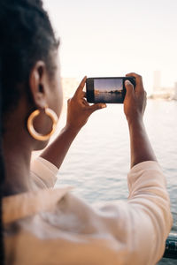 Cropped image of young woman photographing river through smart phone in city