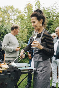 Smiling senior woman holding drink while barbecuing by friends in back yard