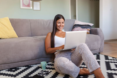 Smiling woman using laptop while sitting by sofa at home