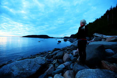 Rear view of woman standing on rock by sea against sky during sunset