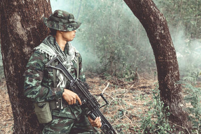 Male army soldier holding gun while standing in forest
