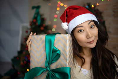 Pretty asian girl in santa hat holding gift box looking at camera.