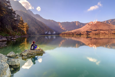 Side view of man fishing in lake while sitting on rock against sky