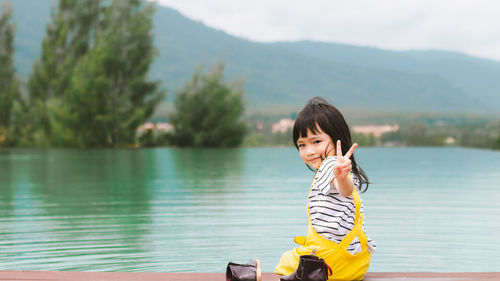 Portrait of cute gesturing peace sign while sitting on pier over lake