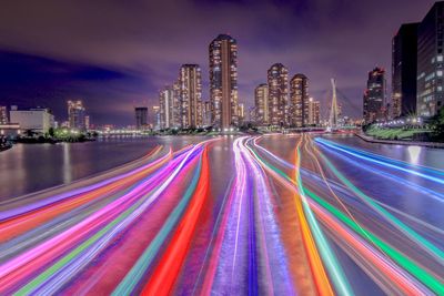 Light trails on road by illuminated buildings against sky at night
