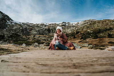 Woman wearing knit hat sitting on footpath with dog against mountains