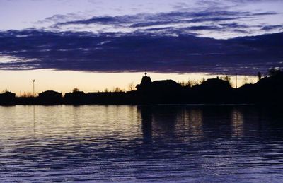 Silhouette buildings by lake against sky during sunset