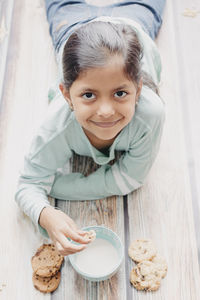 High angle portrait of cute smiling girl eating cookie while lying on hardwood floor