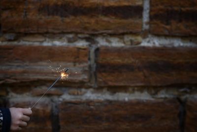 Cropped hand holding lit sparkler by brick wall