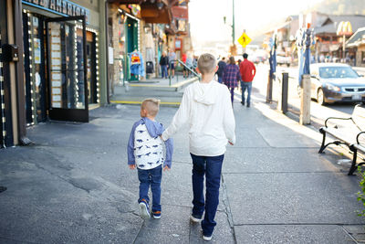 Rear view full length of boy walking with brother on sidewalk in city