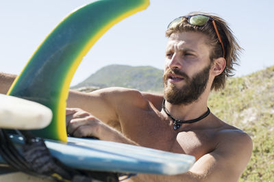 Close-up of man taking surfboard from car roof against clear sky during sunny day