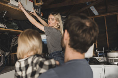 Mid adult woman arranging box on shelf while looking at family in storage room