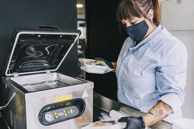 Female chef using airtight packing machinery while holding take out food at restaurant during pandemic