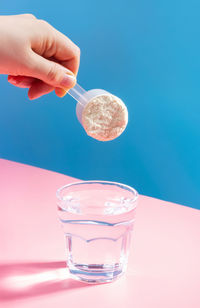 Female hand pouring protein powder into a glass of water on a blue pink background.