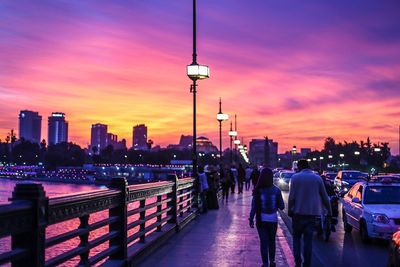 People walking in illuminated city against sky during sunset