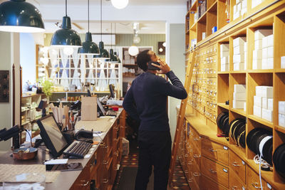 Confident young male owner talking through mobile phone while looking up at rack in furniture store