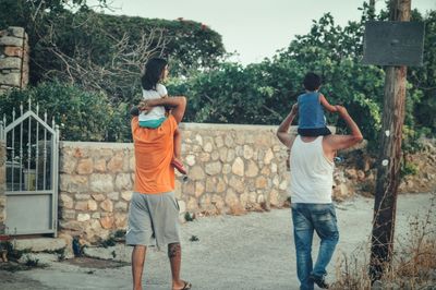 Rear view of men carrying children on shoulder while walking on walkway
