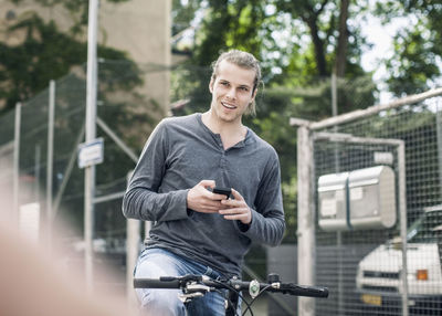 Smiling young man holding mobile phone sitting while on bicycle at street