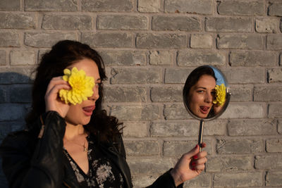 Woman holding yellow flower and mirror with reflection against brick wall