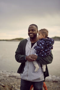 Smiling man carrying boy at beach against sky